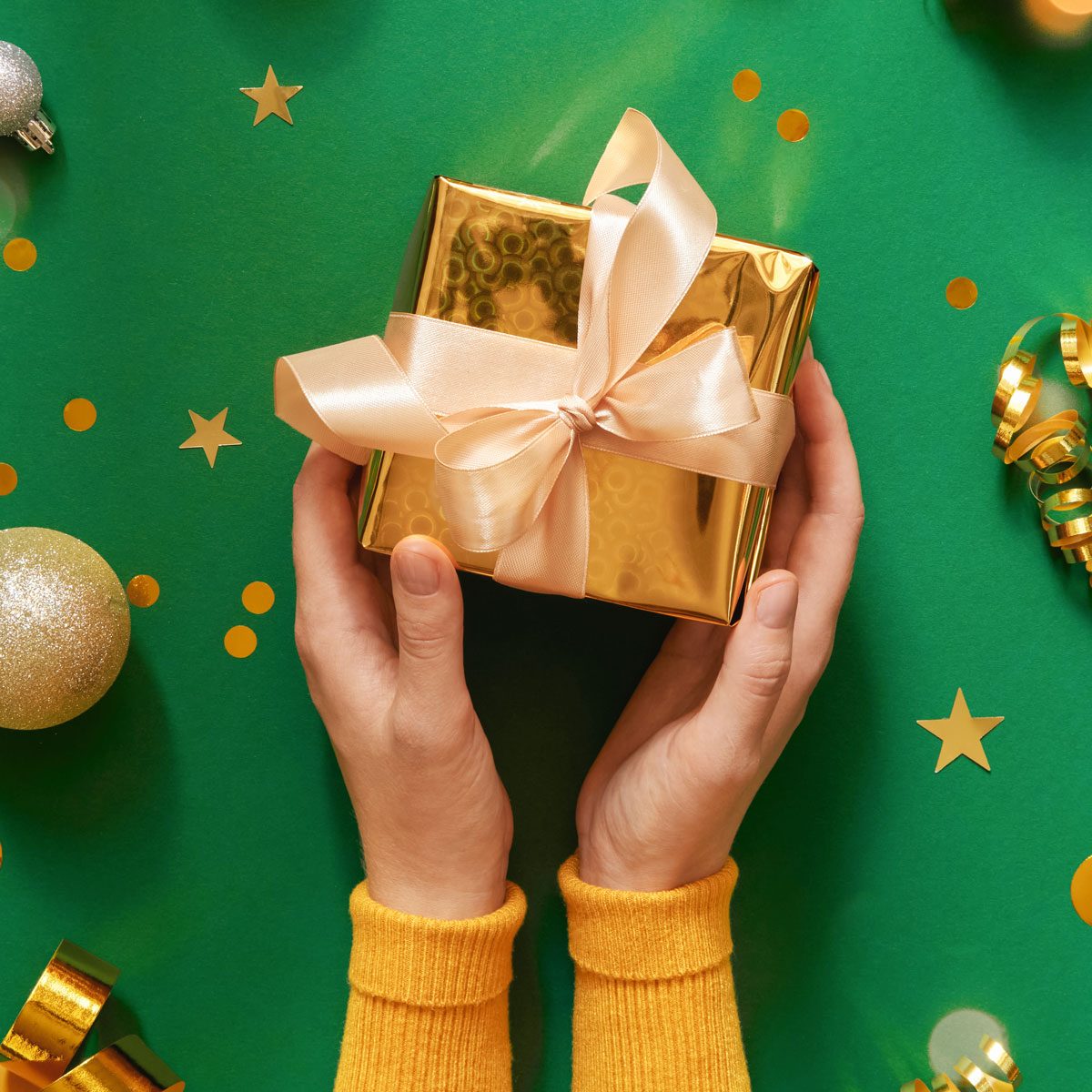 Festive Holiday Background. Hands Holding Gift Box Wrapped In Golden Paper. Decorative Golden And Glittering Ornaments On A Green Backdrop. Top View, Flat Lay