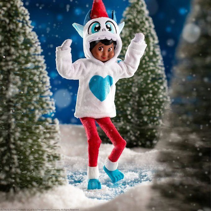 A dark-skinned Elf on the Shelf doll dressed in a white hoodie with a blue heart, red pants, and a blue-and-red winter hat stands in a snowy, pine tree-filled winter scene. Snow is falling gently around the elf.