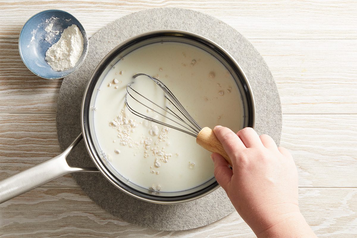 A hand holding a whisk stirring flour into milk in a saucepan, with a small blue bowl of flour nearby on a light wooden surface.