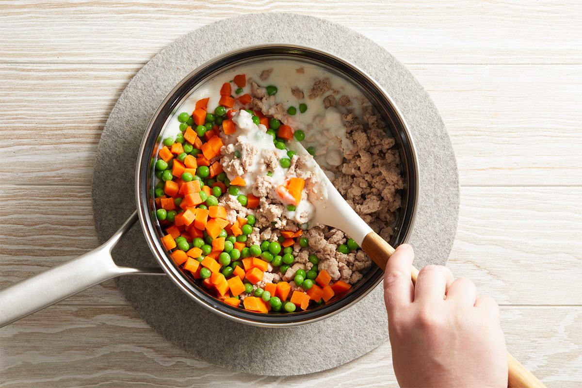 A hand stirs a pot containing ground meat, diced carrots, peas, and a creamy white sauce with a white spatula, on a light-colored wooden surface.
