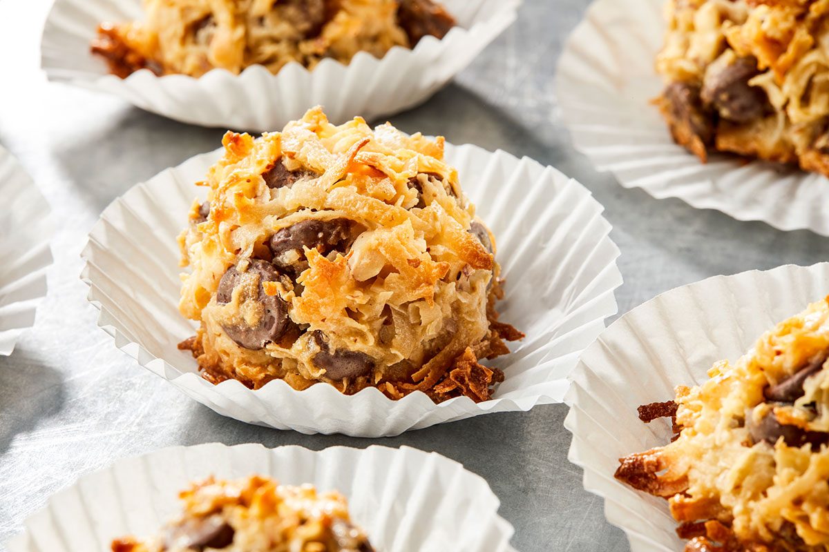 Overhead shot of several Almond Joy Cookies with chocolate chips in white paper liners, arranged on a gray surface;