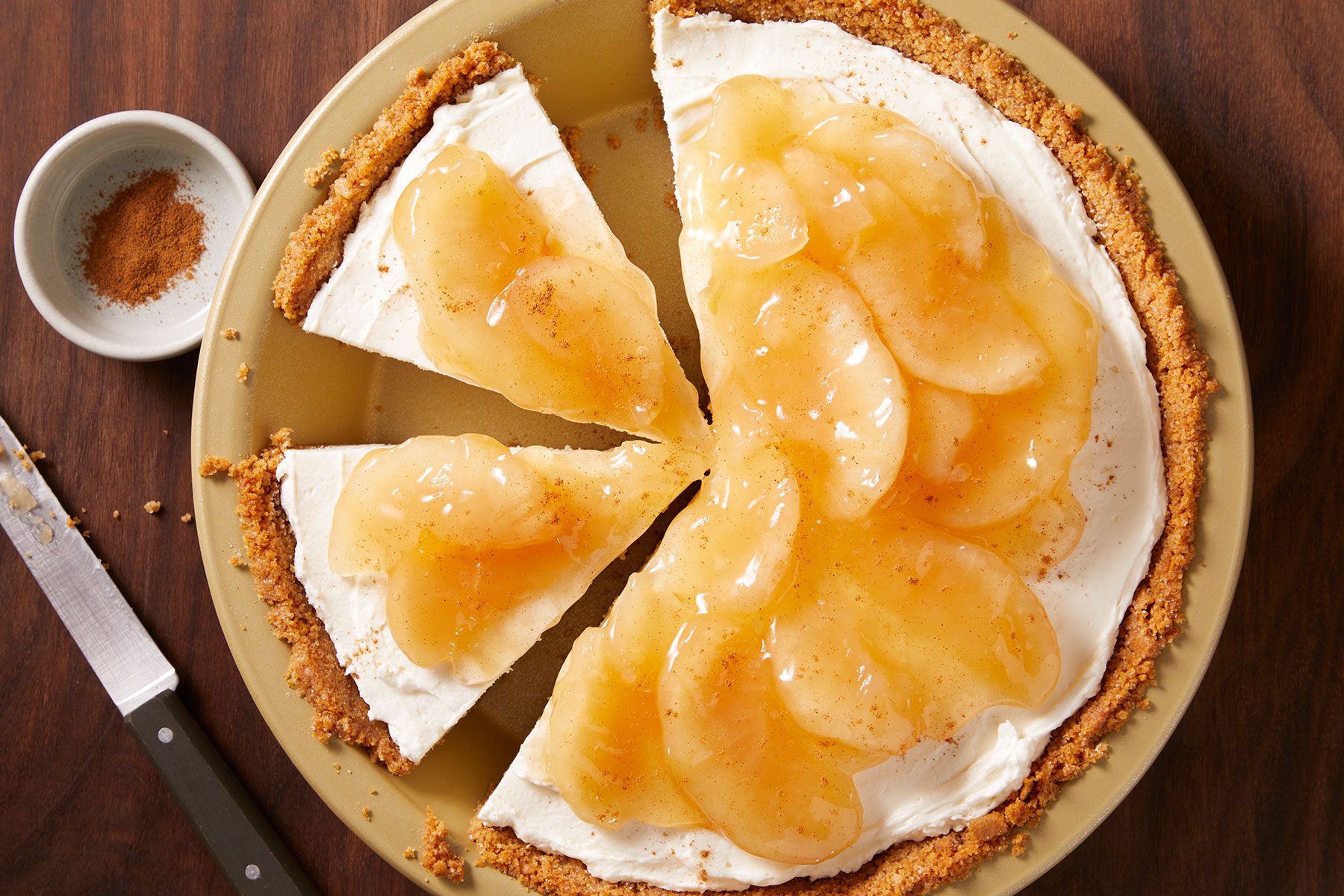 Apple Cream Cheese Pie on a plate cut into few pieces with a knife placed on side and a small bowl of cinnamon. 