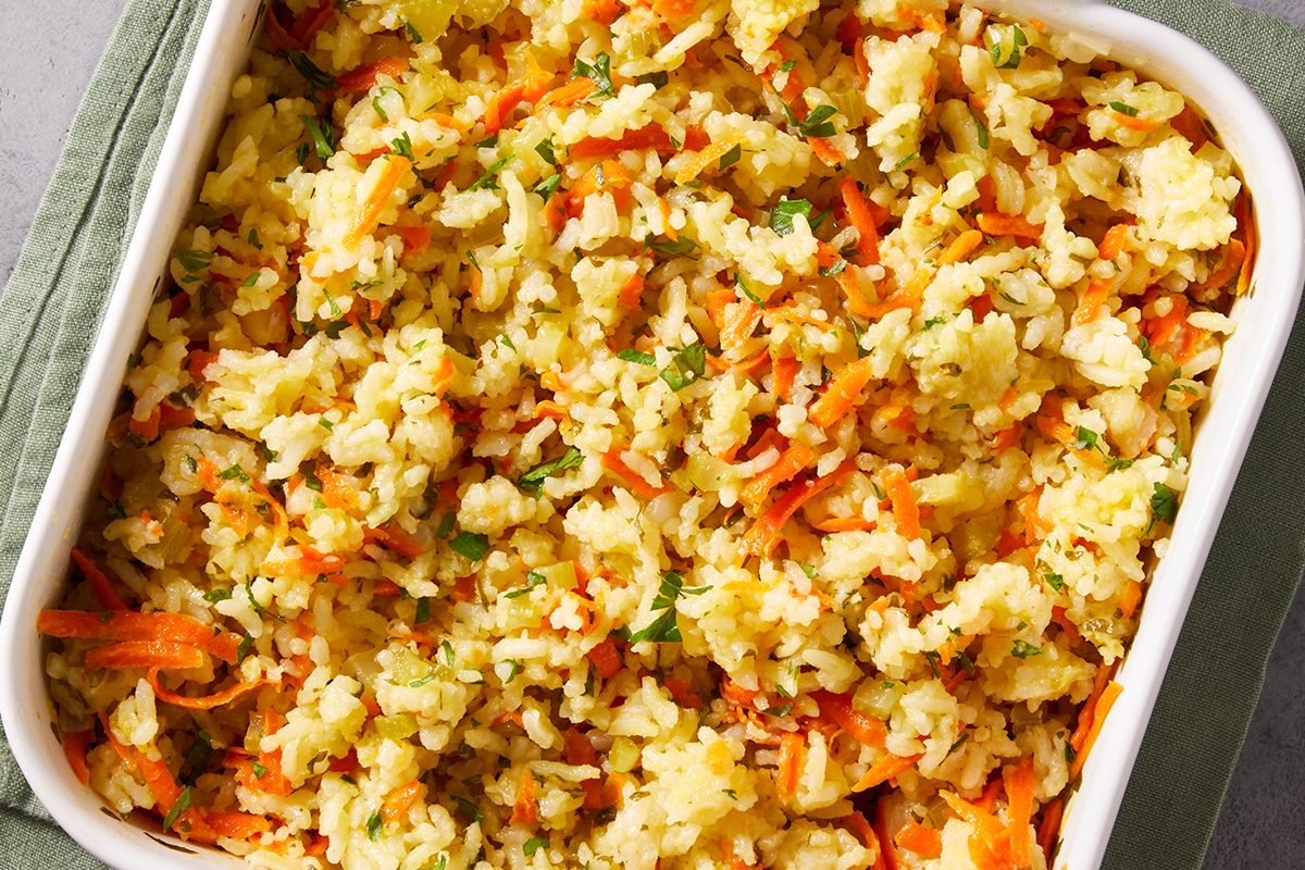 Vertical shot of a white baking dish filled with Baked Rice Pilaf with vegetables, resting on a green napkin with a serving spoon beside it.