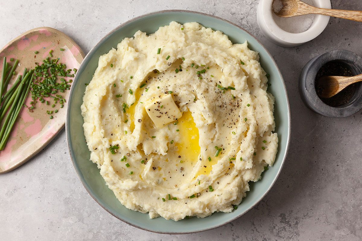 Overhead shot of Boursin Mashed Potatoes served in a wide bowl, swirled creamy and topped with a melting pat of butter, cracked black pepper, and chopped chives; On the side sits a small plate with snipped chives, along with a salt cellar and pepper grinder on a light gray surface;