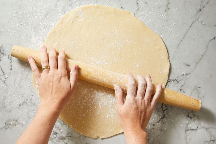 A pair of hands rolling out pie dough with a wooden rolling pin on a floured marble surface.