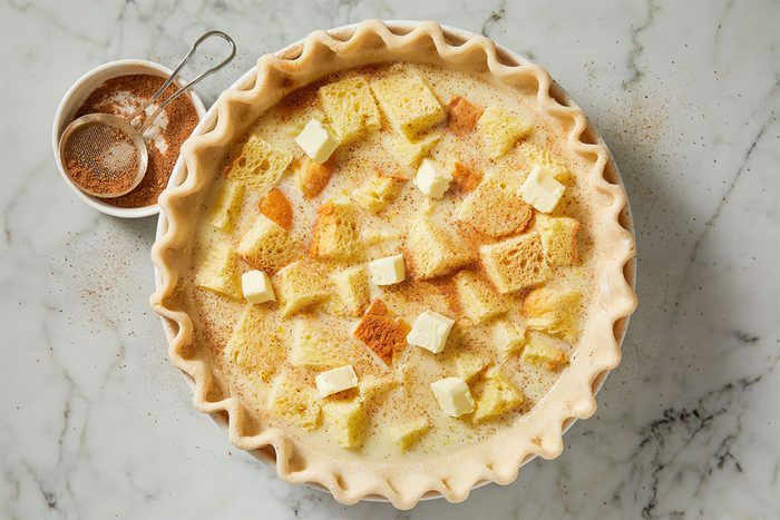A pie crust filled with a custard mixture, topped with cubed bread and small pieces of butter, is shown on a marble surface next to a bowl of ground spices and a small sifter.