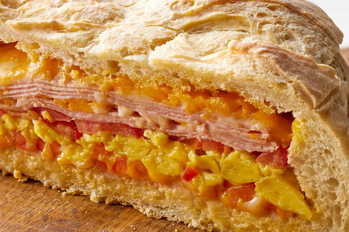 Closeup shot of a sliced breakfast loaf showing layers of scrambled eggs, melted cheese, diced peppers, and thinly sliced ham encased in a golden, flaky bread crust;