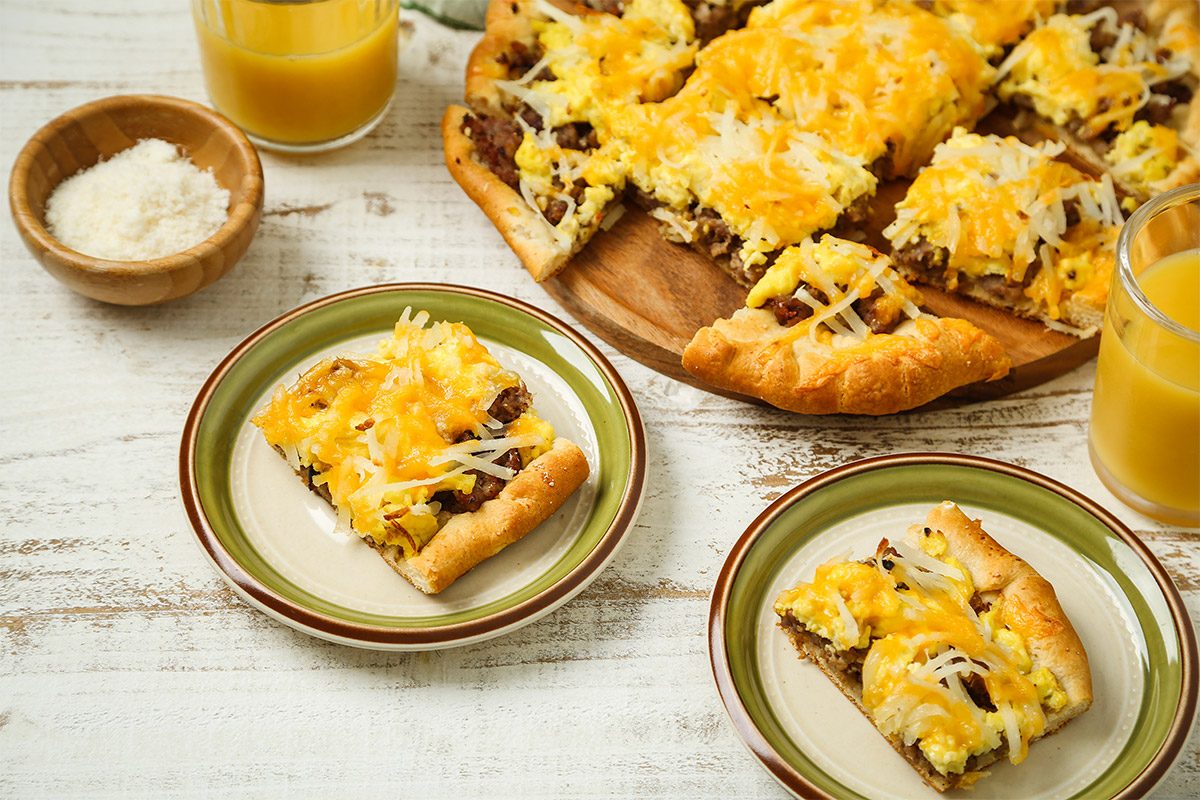 High angle view shot of Brunch Pizza; cut into wedges and placed on a wooden tray; Portions are served on green plates alongside glasses of a refreshing drink; A napkin lies nearby, all arranged on a white and brown wooden surface;