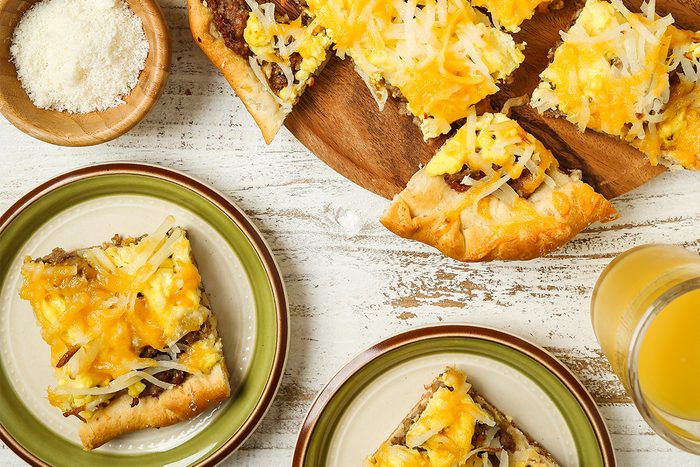 Top view shot of Brunch Pizza; cut into wedges and placed on a wooden tray; Portions are served on green plates alongside glasses of a refreshing drink; A napkin lies nearby, all arranged on a white and brown wooden surface;