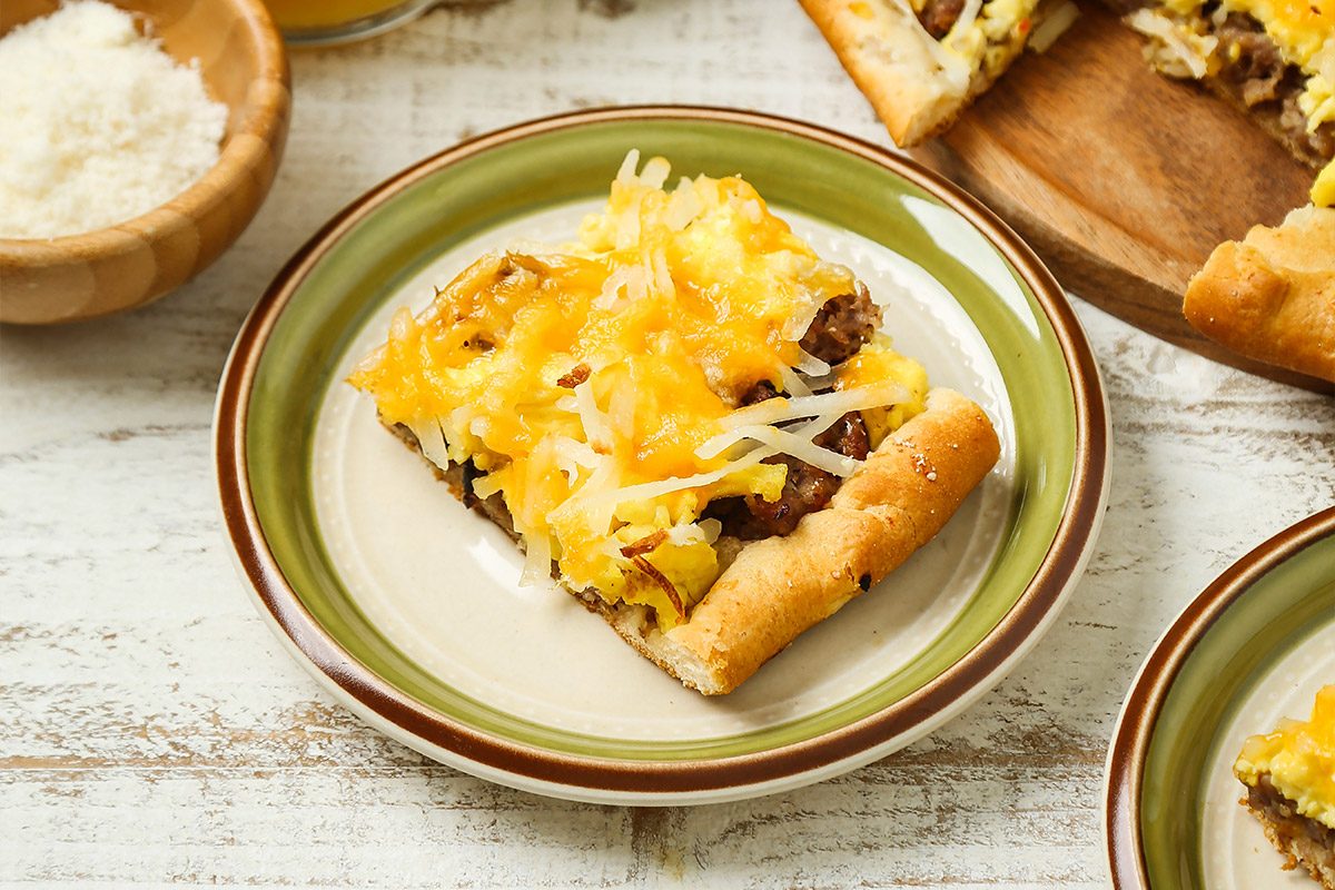 3/4 angle view shot of Brunch Pizza; cut into wedges and placed on a wooden tray; Portions are served on green plates alongside glasses of a refreshing drink; A napkin lies nearby, all arranged on a white and brown wooden surface;