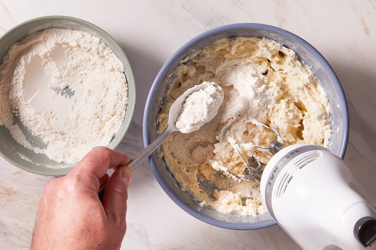 Overhead shot of flour being added with a spoon into a creamed butter mixture in a blue bowl with a hand mixer.