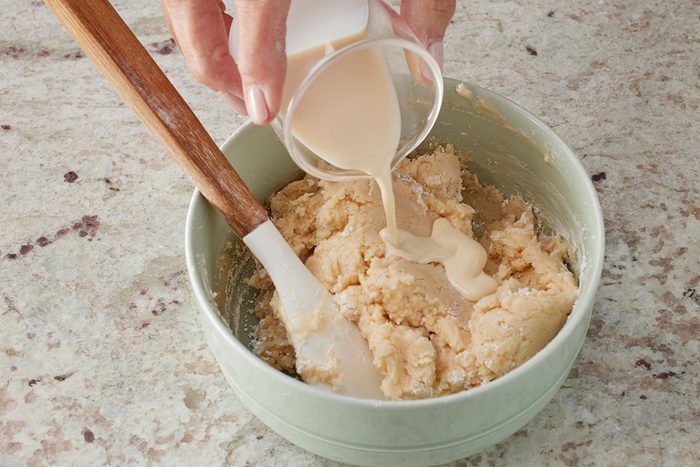 Close shot of a hand pouring liquid from a small cup into a bowl of dough or batter, with a spatula resting inside the bowl on a light-colored countertop;