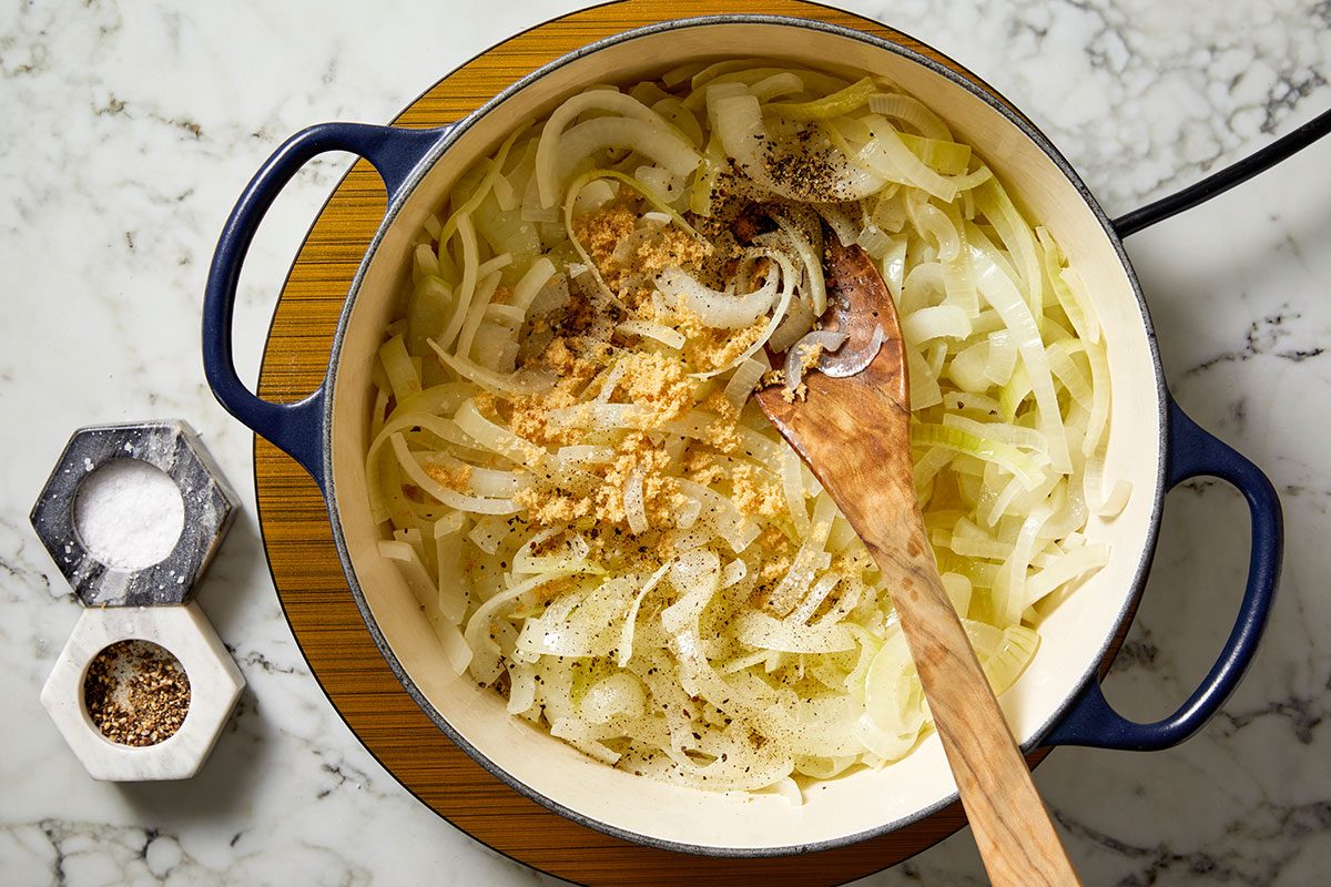 Overhead shot of a pot with sliced onions being sautéed and seasoned with salt and pepper, stirred with a wooden spoon, with salt and pepper containers nearby on a marble countertop.