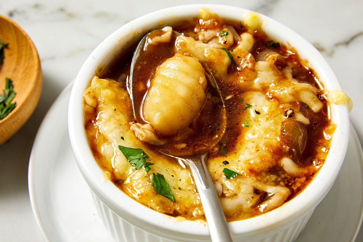 Closeup shot of a bowl of French Onion Gnocchi Soup topped with melted cheese, with a spoon lifting a piece of gnocchi; Fresh parsley is placed nearby.