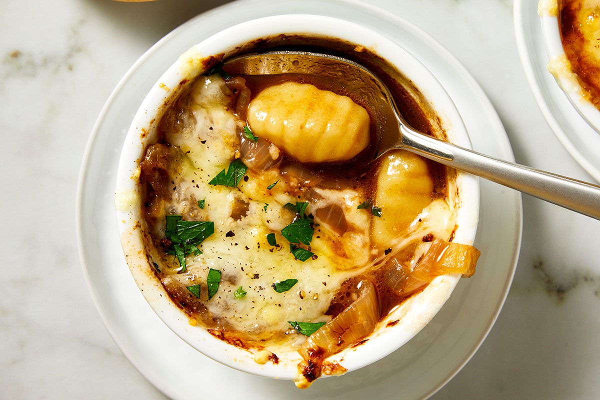 Closeup shot of a bowl of French Onion Gnocchi Soup topped with melted cheese, with a spoon lifting a piece of gnocchi; Fresh parsley is placed nearby;