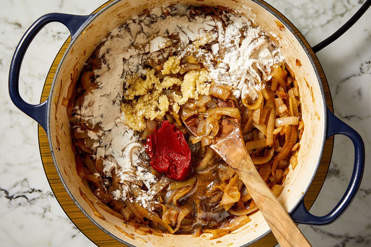 Overhead shot of a blue Dutch oven on a marble countertop containing sautéed onions, minced garlic, flour, and a spoonful of tomato paste being stirred with a wooden spoon.