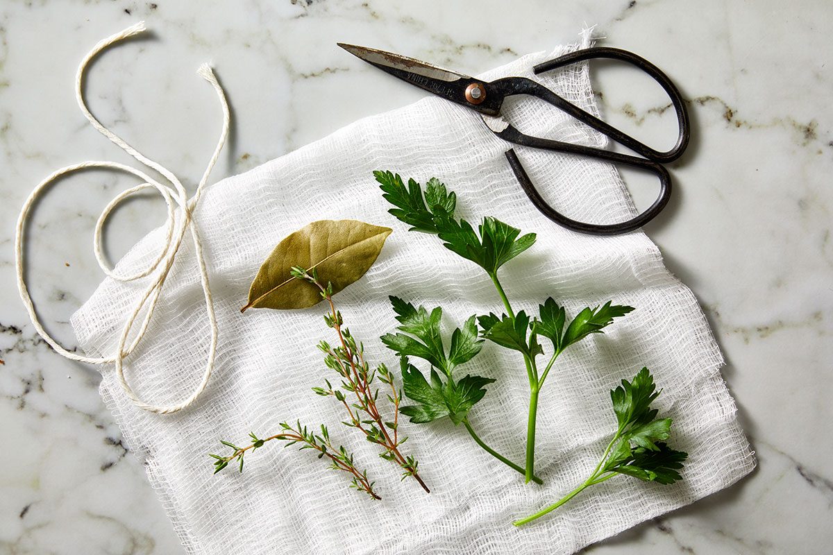 Overhead shot of a bundle of fresh herbs (bay leaf, thyme, and parsley) laid on white cheesecloth, with kitchen twine and a pair of black scissors on a marble surface;