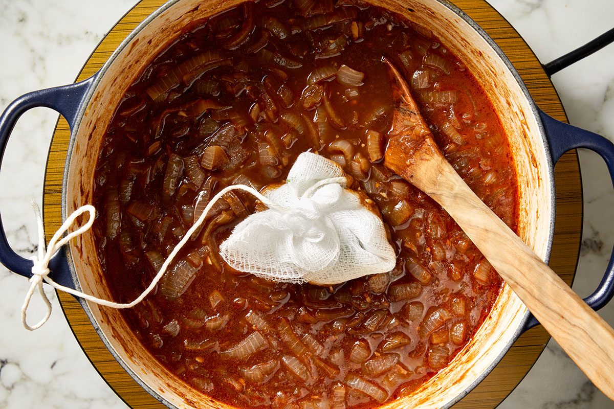 Overhead shot of a pot of rich red stew with chopped onions and a bundle of herbs wrapped in cheesecloth tied with string resting on top, with a wooden spoon inside; The pot sits on a marble countertop.