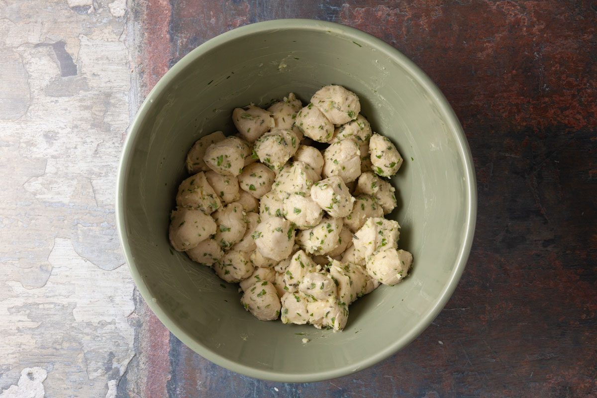 Overhead shot of a green bowl filled with small round dough balls mixed with chopped herbs, resting on a rustic, textured surface.