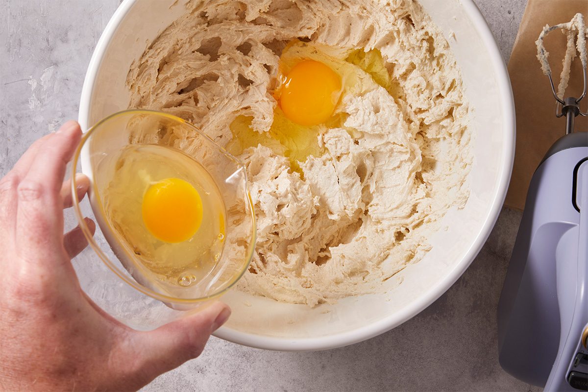 Overhead shot of eggs being added into a mixing bowl filled with creamed butter and sugar for cookie dough preparation.
