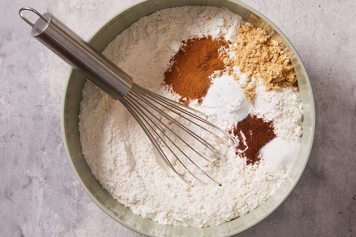 Overhead shot of dry ingredients, flour, ginger, cinnamon, and baking powder combined in a mixing bowl with a whisk.