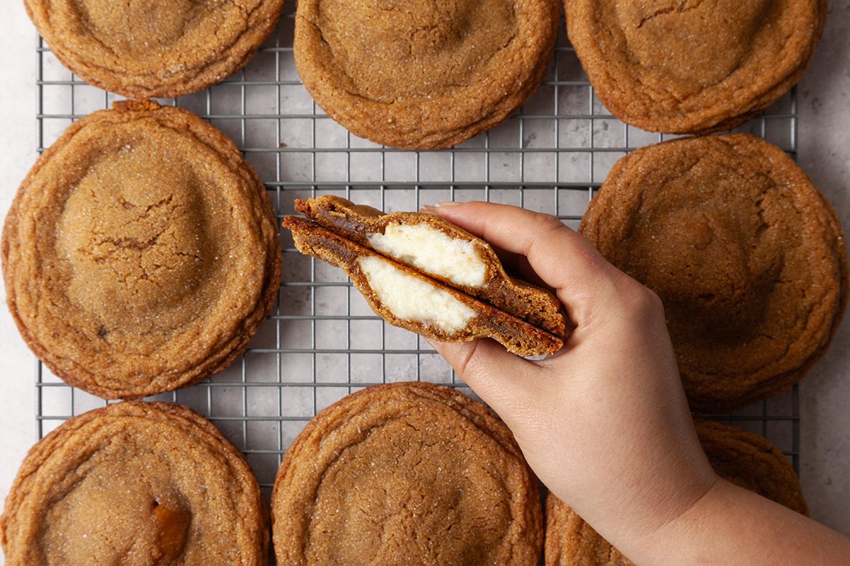 Overhead shot of Gingerbread Cheesecake Cookies on a wire rack, with a hand holding one cookie broken in half to reveal its creamy cheesecake filling; The golden brown, sugar-coated cookies are arranged neatly around the hand, highlighting their soft centers and spiced edges;