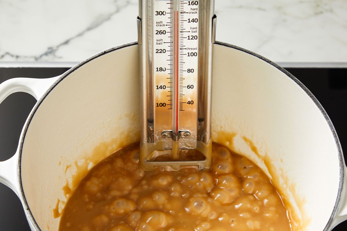 Close-up of boiling caramel mixture in a white pot with a candy thermometer clipped to the side.