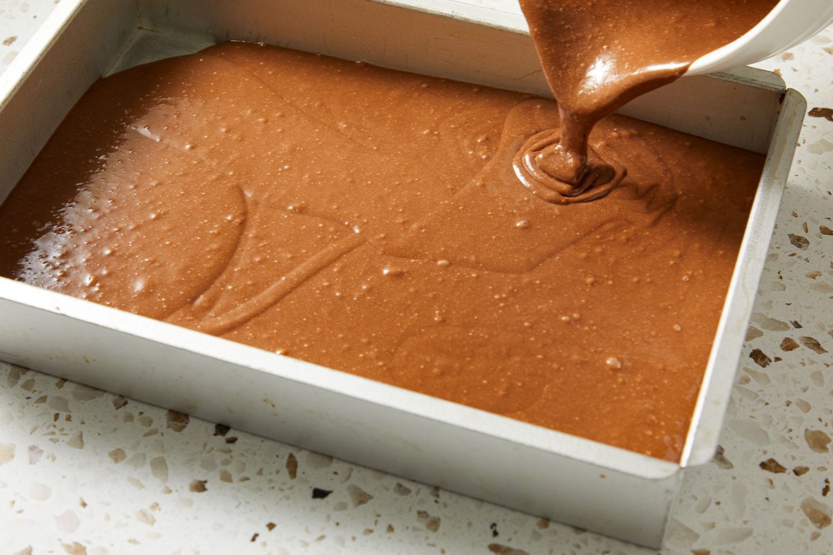 Chocolate cake batter being poured into a rectangular baking pan on a light countertop.