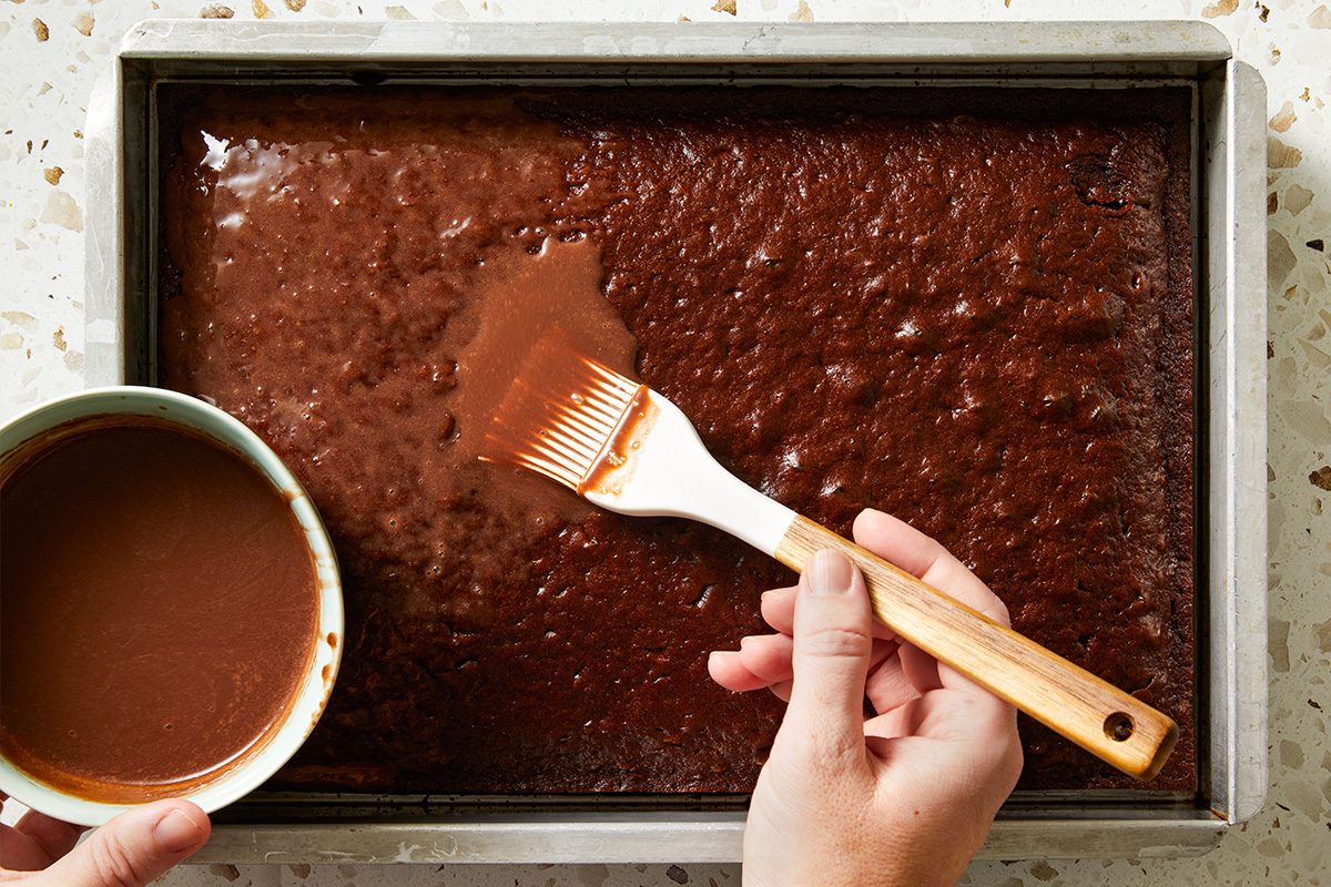 Overhead shot of a baked poke cake in a pan being topped with glossy chocolate sauce using a spatula.
