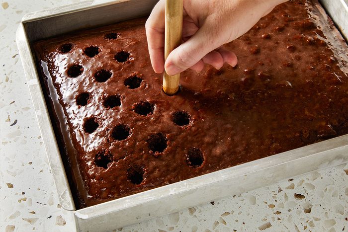 Overhead shot of a baked chocolate cake in a rectangular pan with holes being poked into the surface using the handle of a wooden spoon.