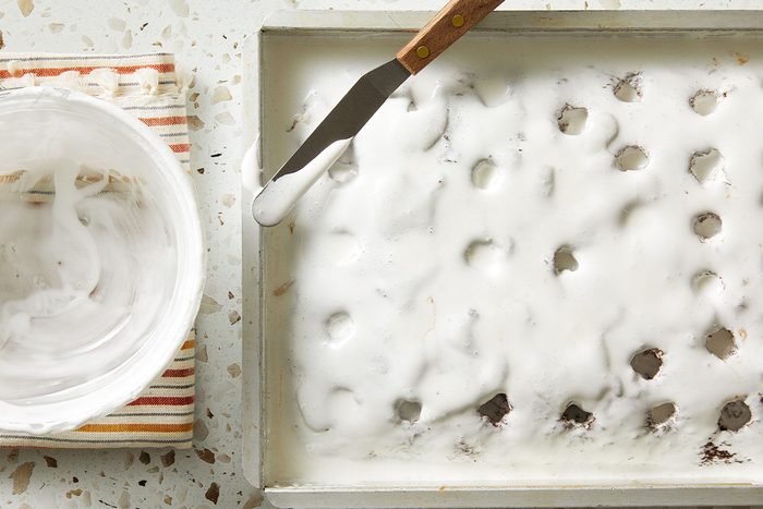 Top-down view of the poke cake covered with a thick layer of whipped cream, with an offset spatula resting nearby.