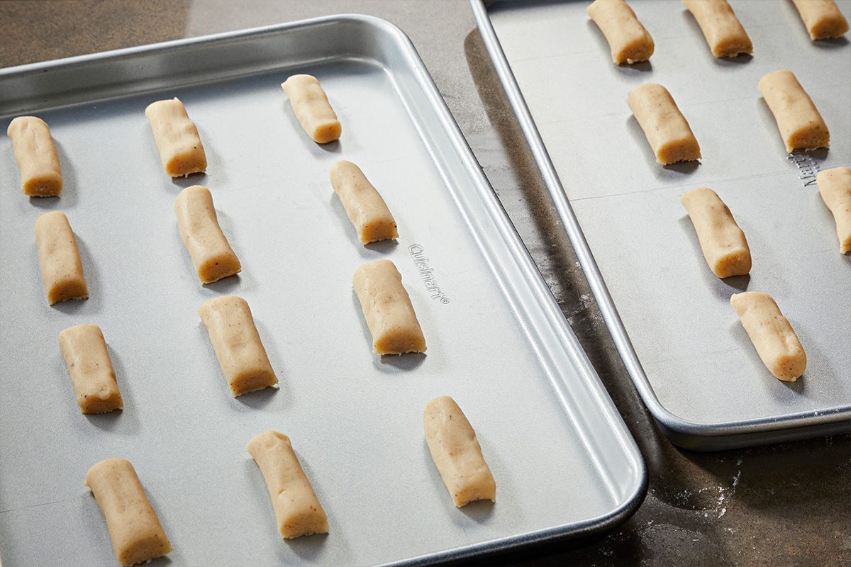 3/4 angle view shot of two baking sheets with evenly spaced, unbaked cookie dough logs arranged in rows, light in color, set on a countertop and ready for baking;