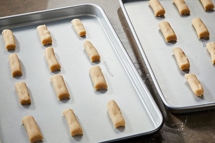 3/4 angle view shot of two baking sheets with evenly spaced, unbaked cookie dough logs arranged in rows, light in color, set on a countertop and ready for baking;