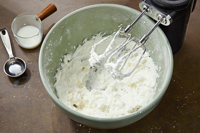 Overhead shot of a mixing bowl filled with thick whipped cream on a counter, with an electric hand mixer resting on the bowl. Nearby are a measuring spoon and a small cup containing liquid;