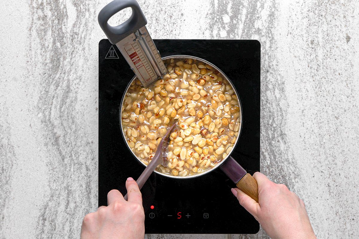 A person stirs a pot of peanuts in liquid on an induction cooktop while monitoring the temperature with a candy thermometer.