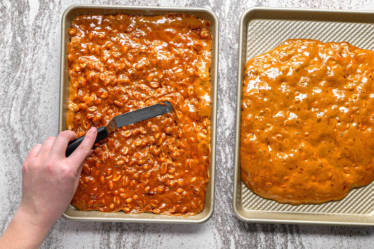 A hand uses an offset spatula to spread peanut brittle mixture evenly on a baking sheet, with another sheet of set peanut brittle next to it on a marble countertop.