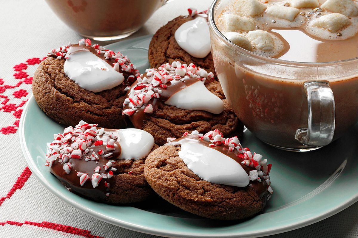 A plate of chocolate cookies topped with white icing and crushed peppermint sits next to a clear mug of hot chocolate with marshmallows.