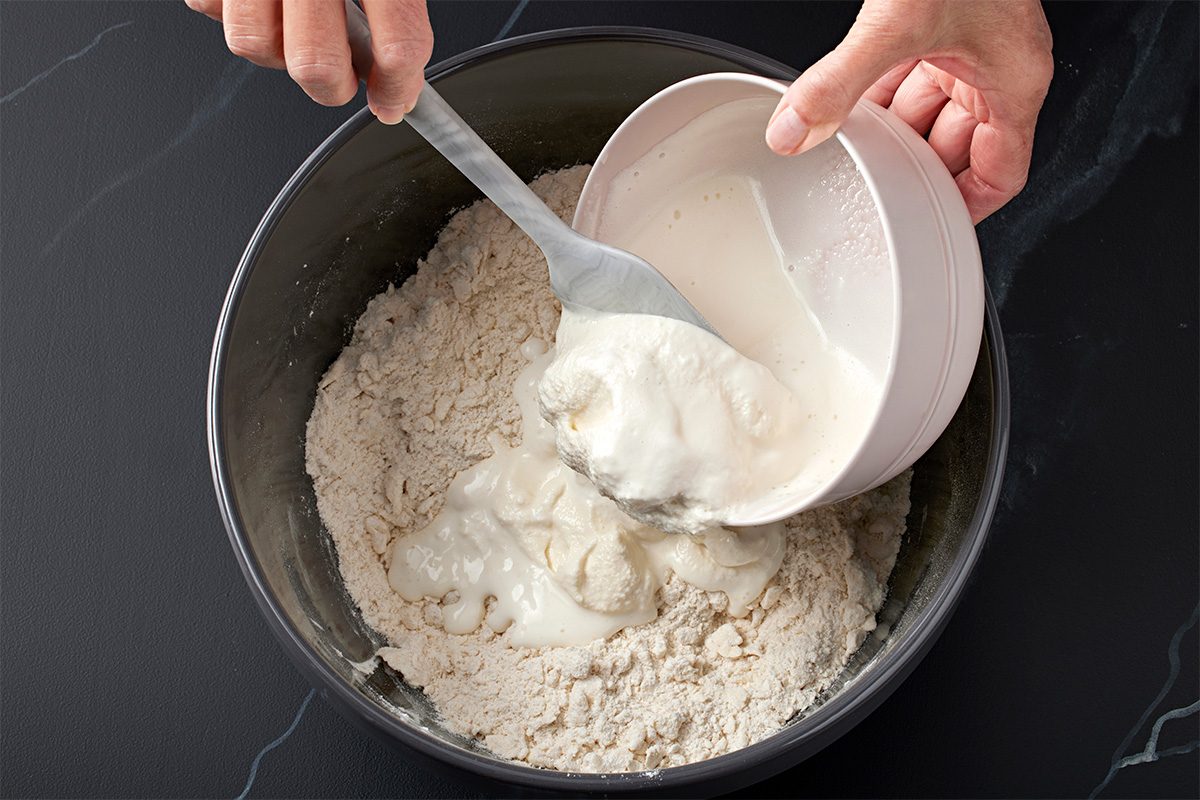 A person pours a bowl of liquid mixture into a larger bowl of flour, stirring with a spoon on a dark countertop.