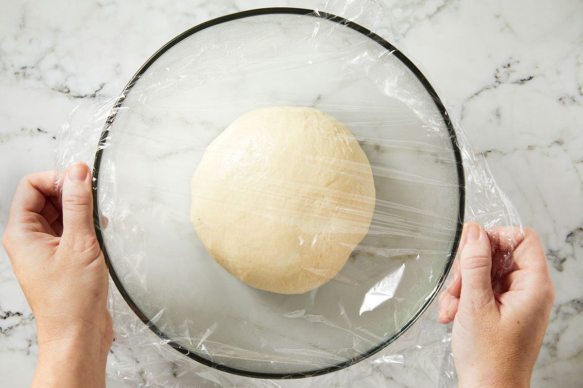 Overhead shot of a pair of hands covering a glass bowl containing a ball of dough with plastic wrap; The bowl rests on a white marble surface. 