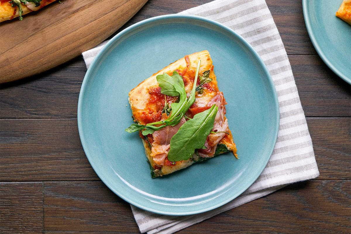overhead shot of A slice of pizza topped with arugula and prosciutto sits on a blue plate, which is placed on a striped cloth on a wooden table; Other pizza slices are visible nearby