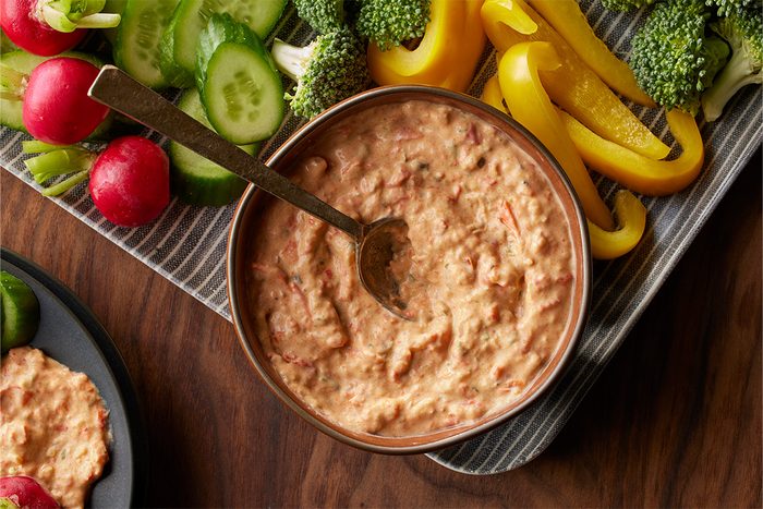 Top-down shot of a bowl of Roasted Vegetable Dip with a spoon, surrounded by cucumbers, radishes, yellow bell pepper strips, and broccoli on a striped tray;
