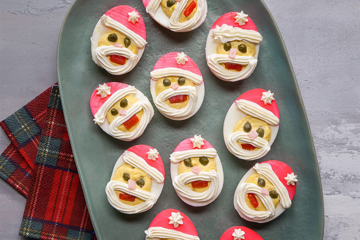 Deviled eggs decorated as Santa Claus faces with red hats, white beards, and candy details, arranged on a dark oval platter next to a red plaid napkin on a gray surface.
