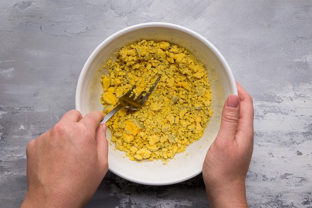A person uses a fork to mash yellow chickpeas in a white bowl, preparing a mixture on a gray textured surface.