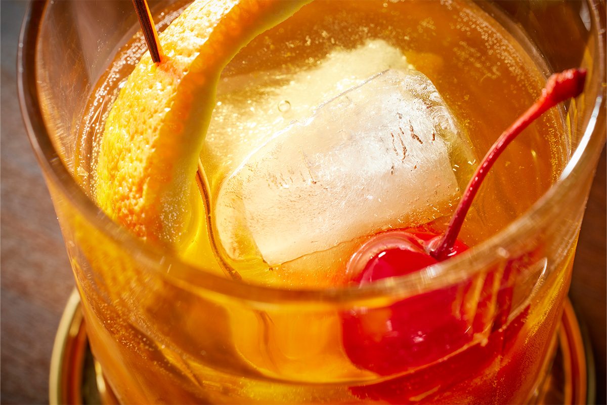 Closeup shot of a Scotch Old-Fashioned in a clear glass, served over a large ice cube, garnished with a twist of orange peel and a maraschino cherry;