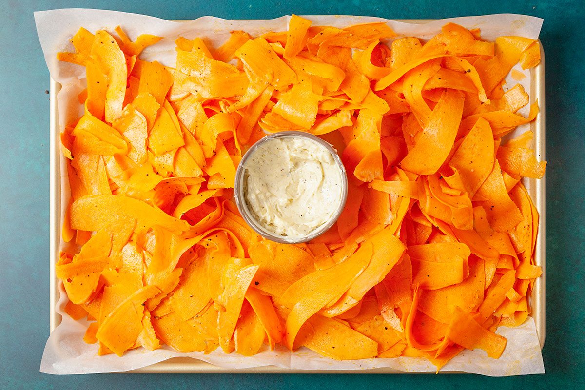 Overhead shot of a tray lined with parchment paper holding thin orange butternut squash ribbons arranged around a small bowl of creamy white dip in the center;