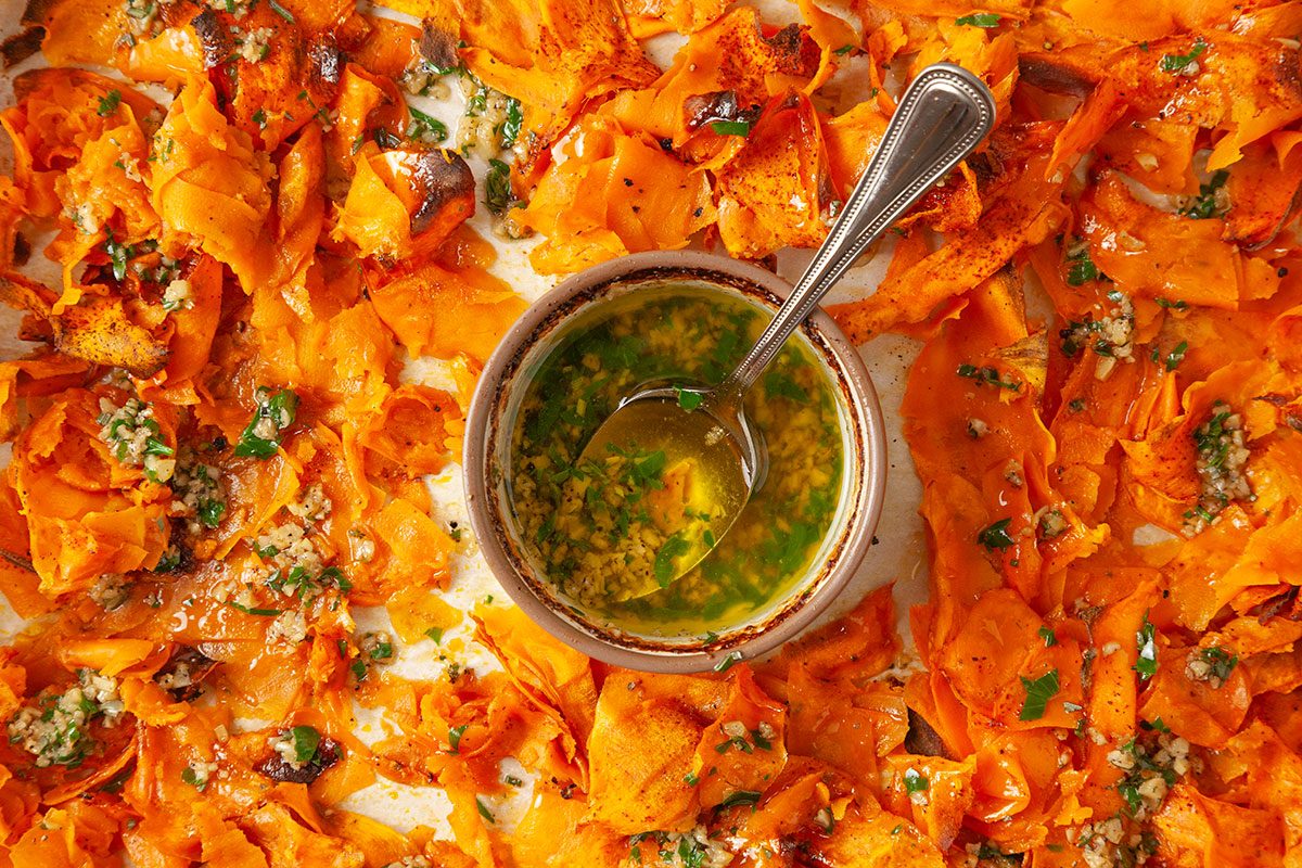 Overhead shot of a bowl of green herb sauce with a spoon on a tray surrounded by roasted, thinly sliced sweet potatoes garnished with herbs and garlic;