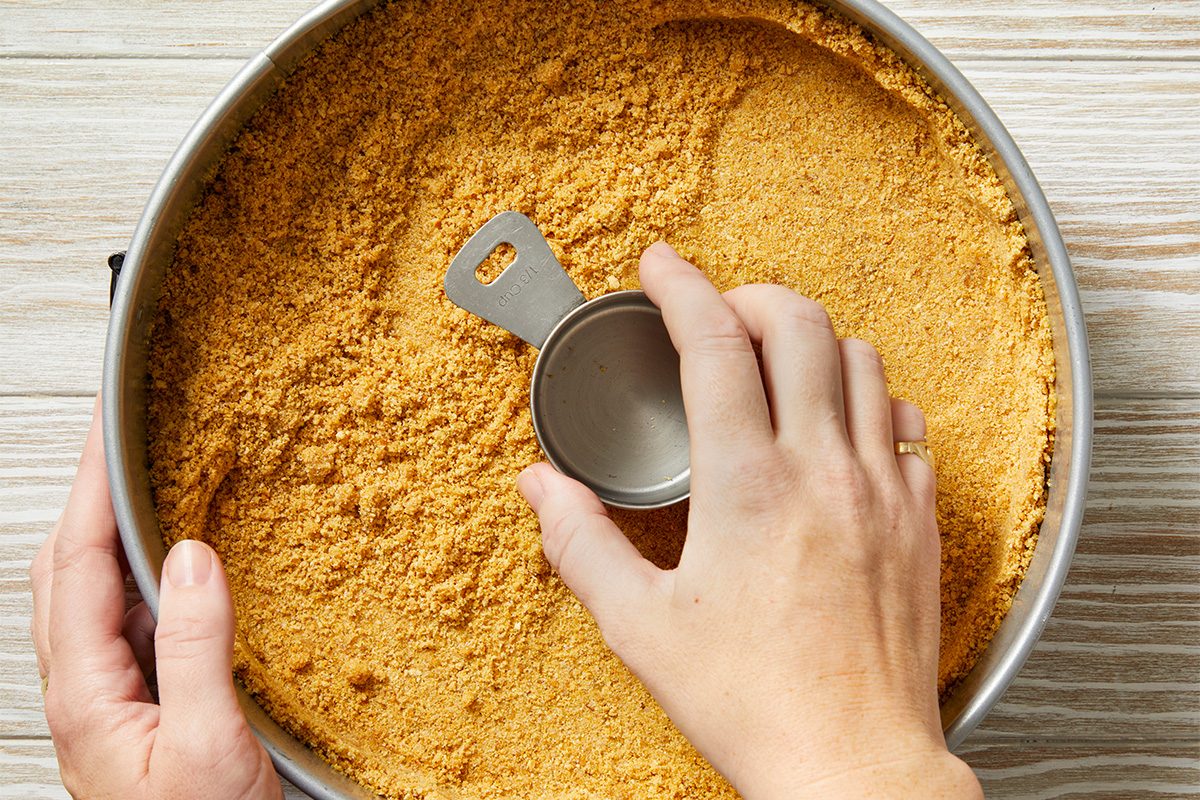 A hand uses a metal measuring cup to press down and smooth a layer of crushed graham crackers in a round pan, preparing a crust on a light wooden surface.