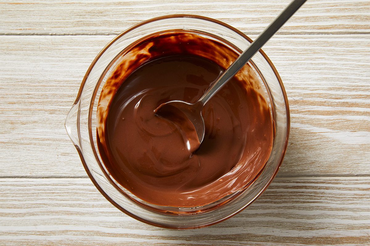 A glass bowl filled with melted chocolate and a metal spoon resting inside, placed on a light wooden surface.
