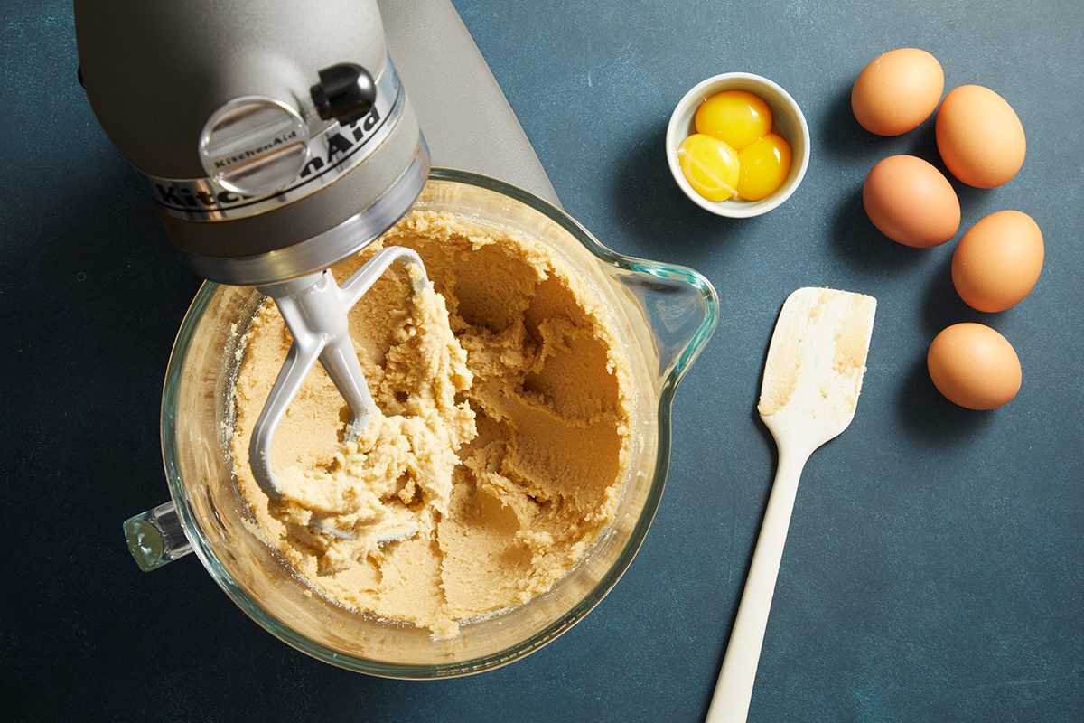 Overhead shot of a stand mixer bowl with creamed butter and sugar inside, surrounded by baking ingredients including eggs and vanilla.