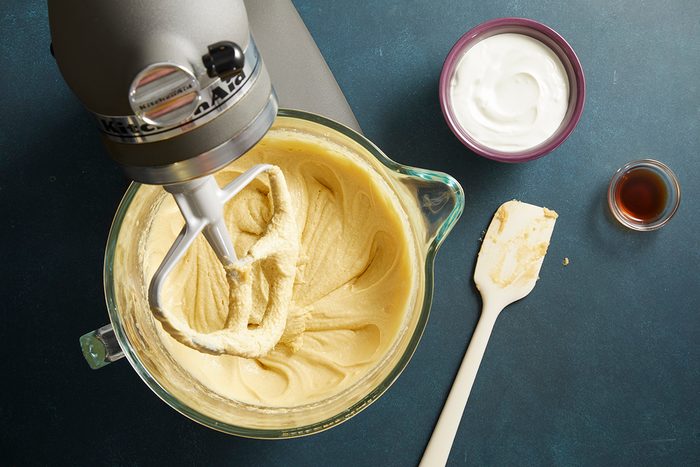 Overhead shot of a stand mixer creaming butter and sugar into a light fluffy batter, spatula placed nearby on a dark surface.